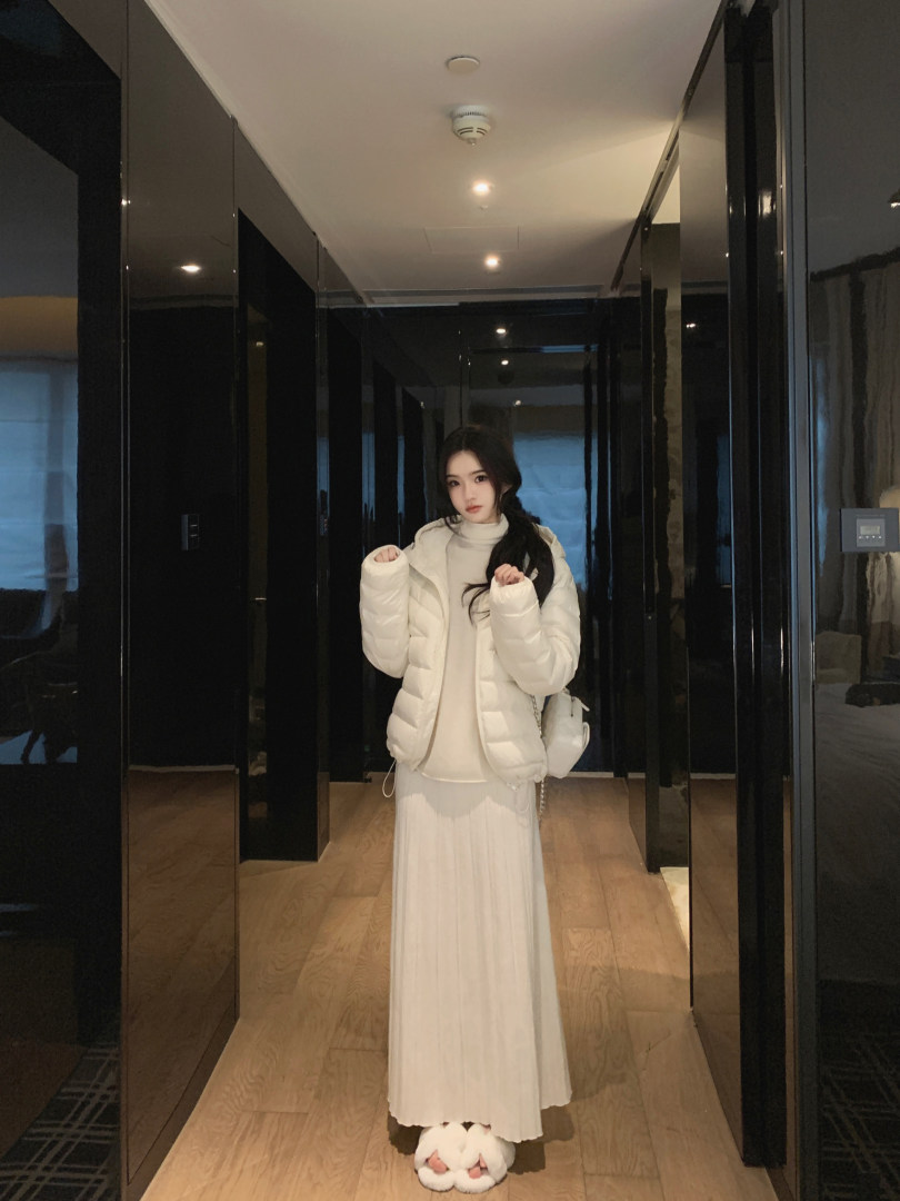 Woman wearing a white puffer jacket, white turtleneck, and pleated skirt in a hallway.