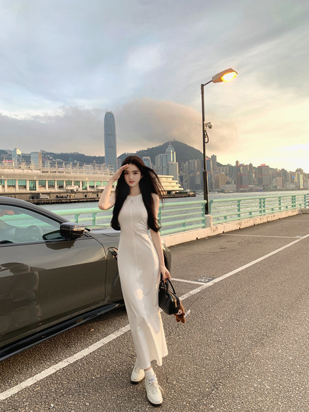 Woman wearing a white sleeveless dress standing next to a car with city skyline in background