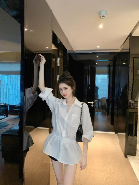 Woman wearing a white shirt dress with rolled sleeves and a black handbag, posing indoors.