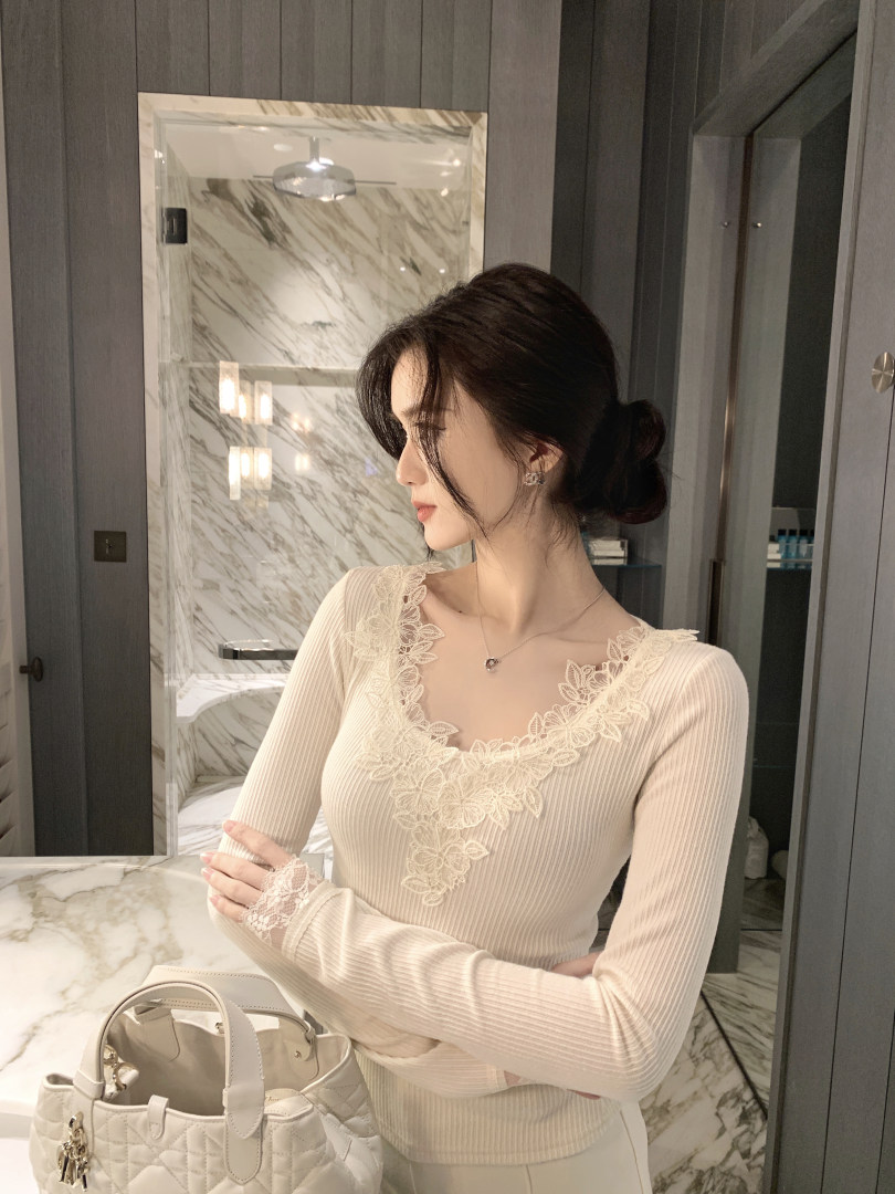 Woman wearing a cream ribbed long-sleeve top with lace floral detailing on neckline and cuffs, standing in a marble bathroom with a white quilted handbag nearby.