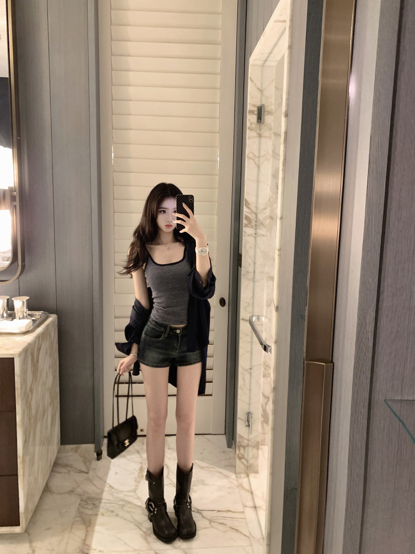 Woman taking mirror selfie wearing striped tank top, denim shorts, and black boots