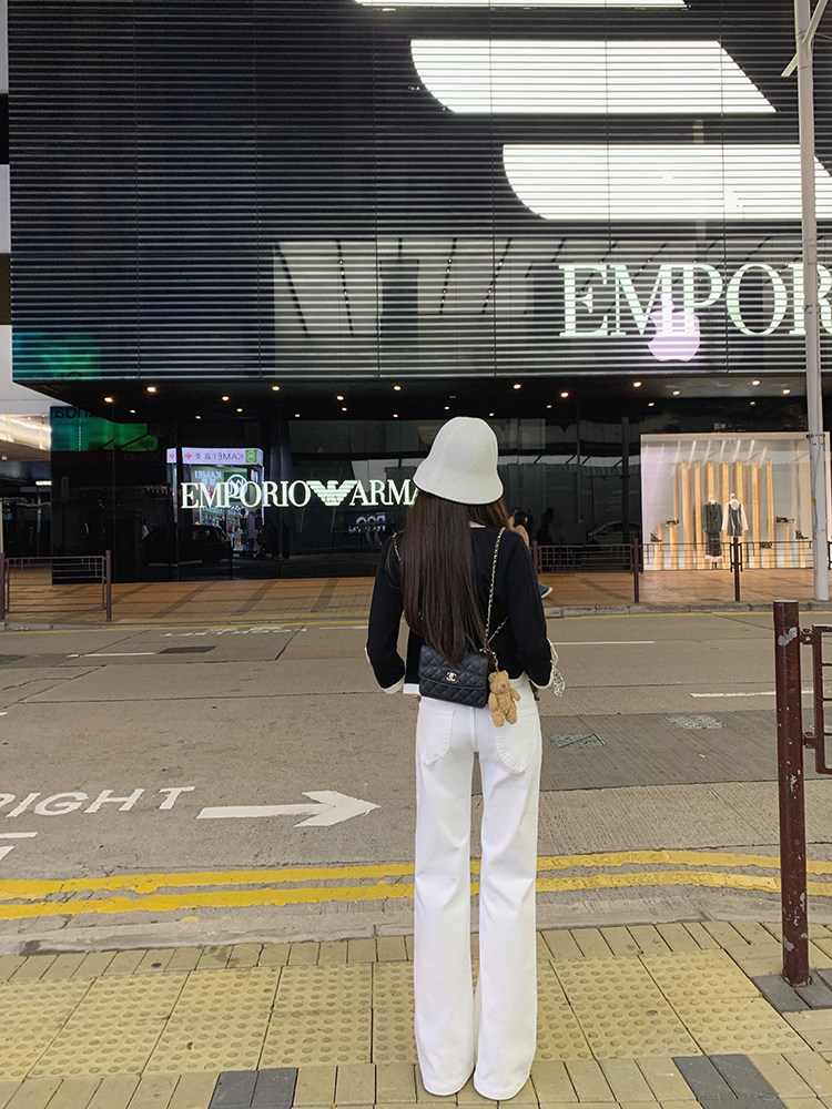 Woman wearing white wide-leg pants, black top, white hat, and black quilted bag, standing on sidewalk in front of Emporio Armani store
