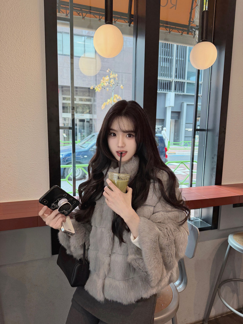 Woman wearing a gray faux fur coat while holding a drink and phone in a cafe setting