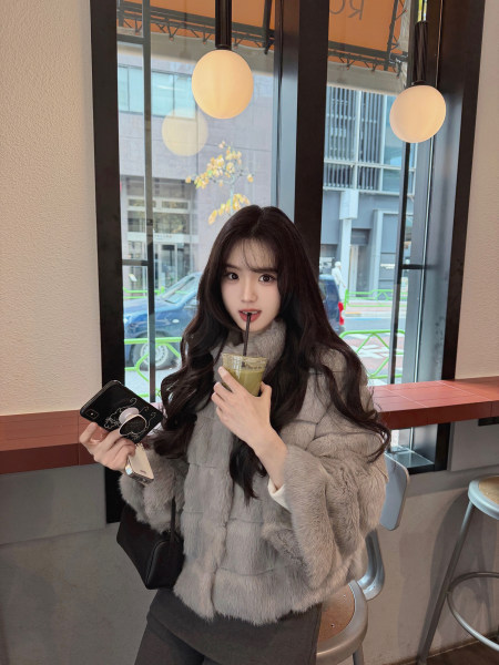 Woman wearing a gray faux fur coat while holding a drink and phone in a cafe setting