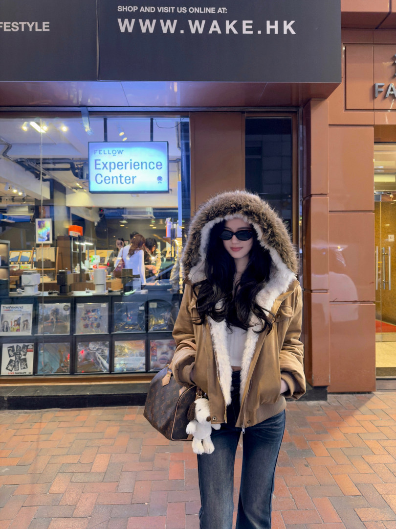 Woman wearing a brown hooded jacket with fur trim, standing outdoors in front of a store.