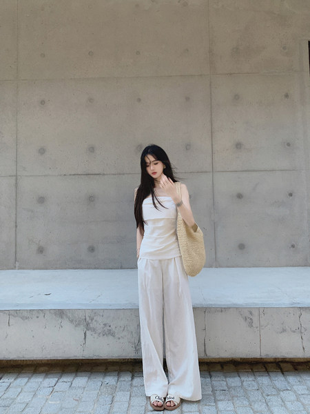 Woman wearing a white sleeveless top and wide-leg pants with a woven bag and sandals.