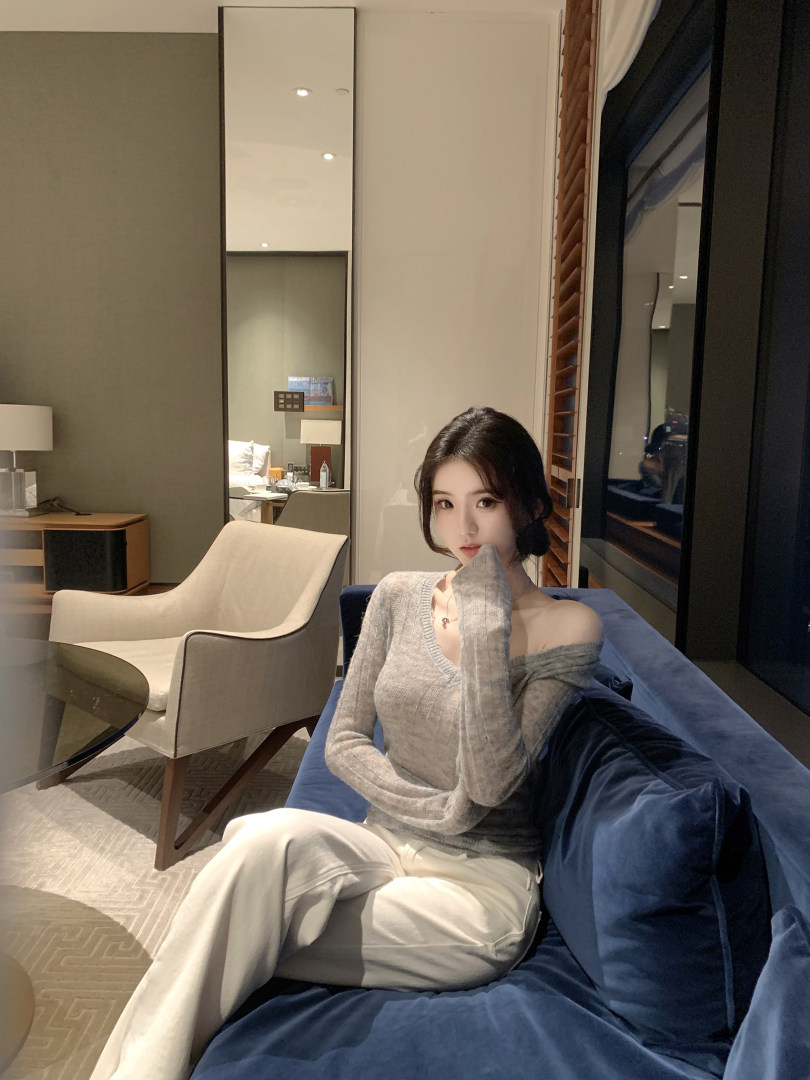 Woman wearing a gray off-shoulder knit top and white pants, sitting on a blue sofa in a modern room