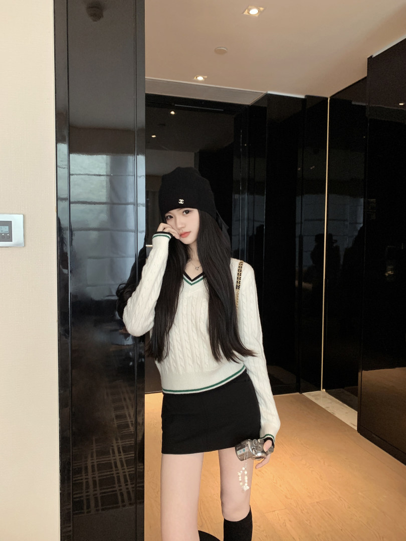 Woman wearing a white cable-knit sweater with green trim, black beanie, and black skirt, holding a camera.