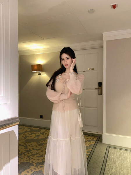 Woman wearing a light pink knit cardigan with floral embellishments and a white tiered tulle skirt, posing in a hotel hallway.