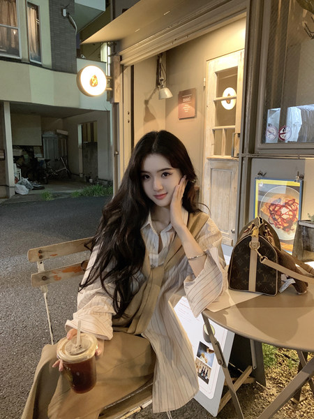 Woman wearing striped shirt and beige vest sitting at outdoor cafe with drink and Louis Vuitton bag