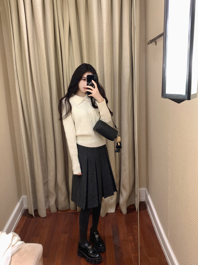 Woman taking mirror selfie wearing cream cable knit sweater, black pleated skirt, black tights, and chunky loafers
