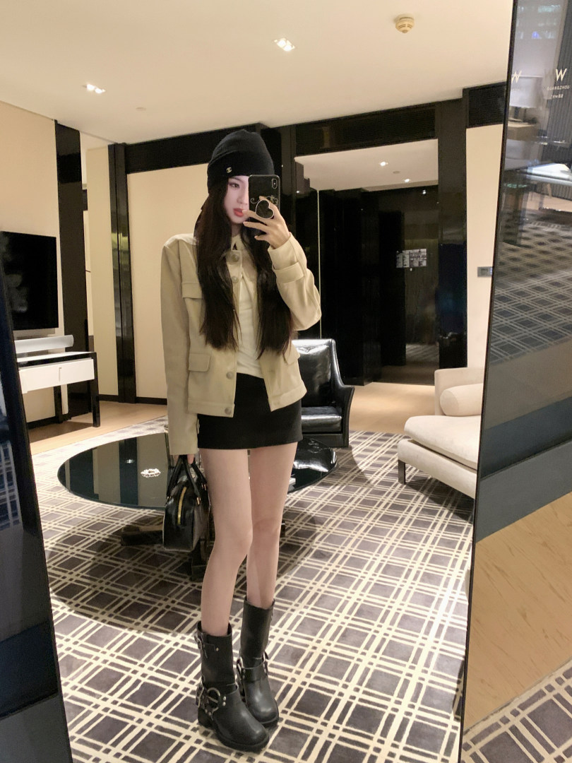 Woman taking mirror selfie wearing beige jacket, black mini skirt, and black boots