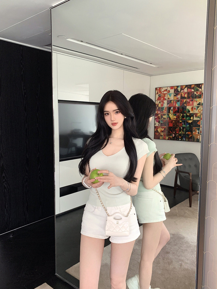 Woman wearing a light green ribbed top and white shorts, holding a green apple, standing in front of a mirror.