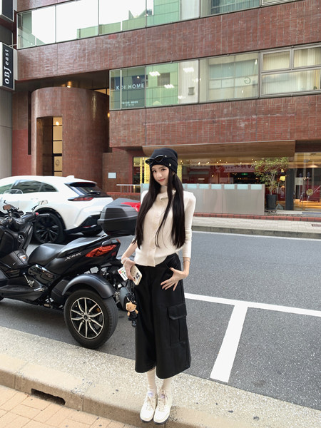 A woman wearing a beige cardigan, black wide-leg pants, and white sneakers standing on a city street.
