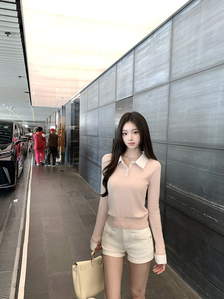 Woman wearing a light pink long-sleeve polo top and cream shorts, holding a beige handbag.