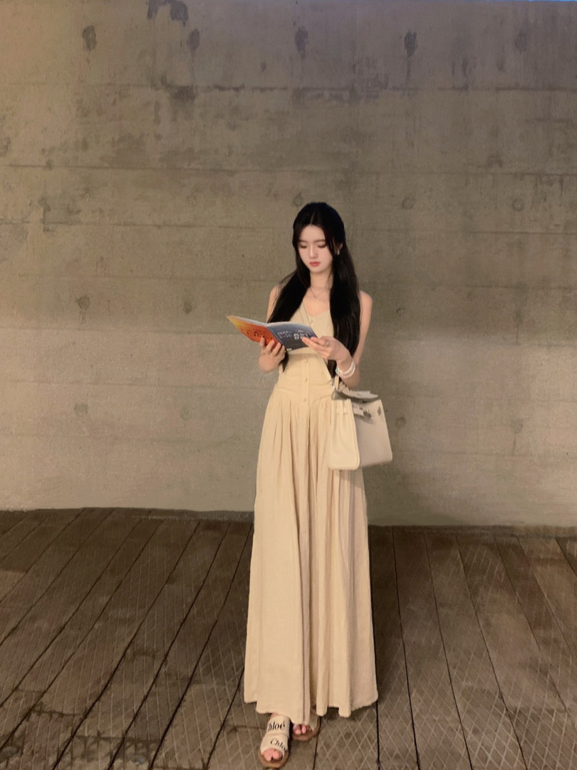 Woman wearing a beige sleeveless long dress with buttons, holding a book, standing against a concrete wall