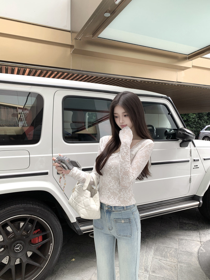 Woman wearing white lace long-sleeve top and light blue jeans, holding camera and white quilted handbag, standing next to white SUV