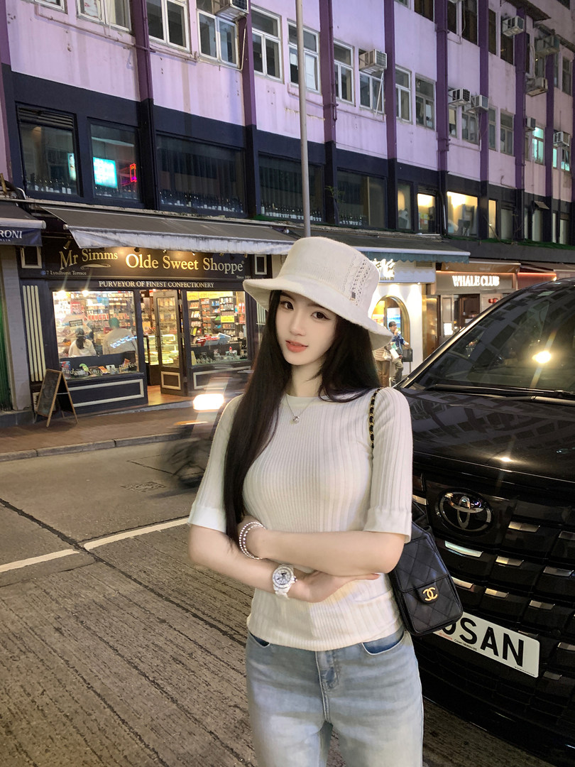 Woman wearing a white ribbed top, light blue jeans, and a white bucket hat, standing on a city street at night with a black quilted handbag.