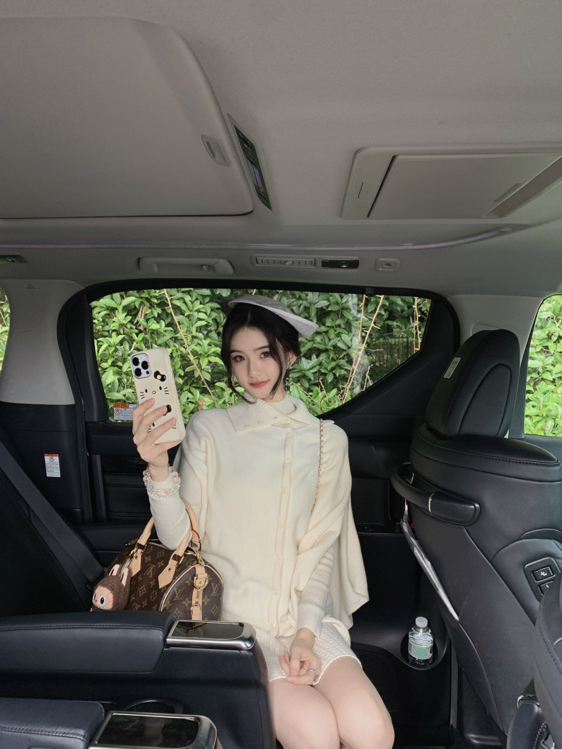 Woman taking a selfie in a car wearing a cream-colored cardigan and hat, with a Louis Vuitton bag beside her.
