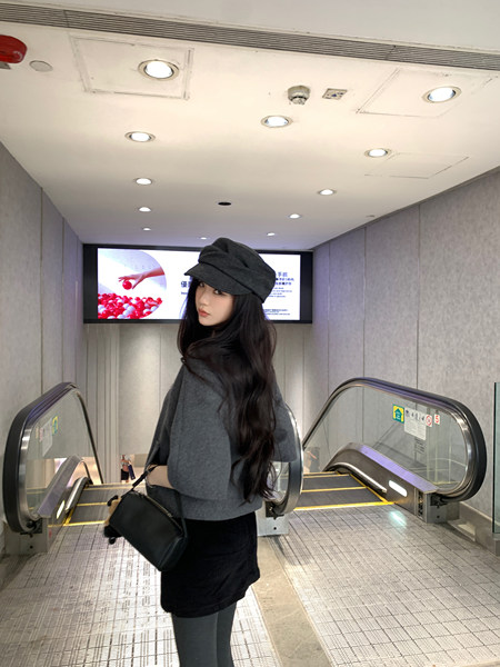 Woman wearing a gray beret, gray sweater, and black skirt standing near escalators.