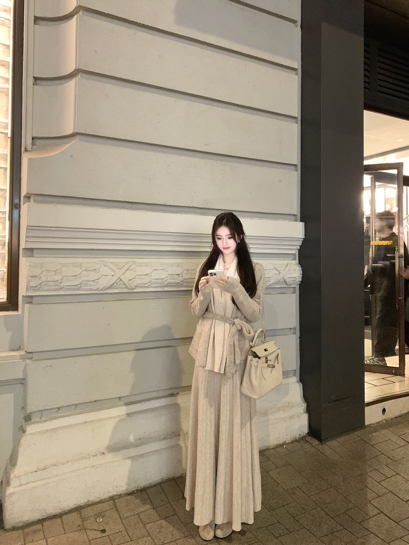 Woman wearing a beige knit cardigan and pleated skirt outfit while holding a phone, standing against a building wall.