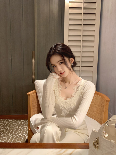 Woman wearing a white lace top and matching pants, seated in a chair with a handbag nearby.