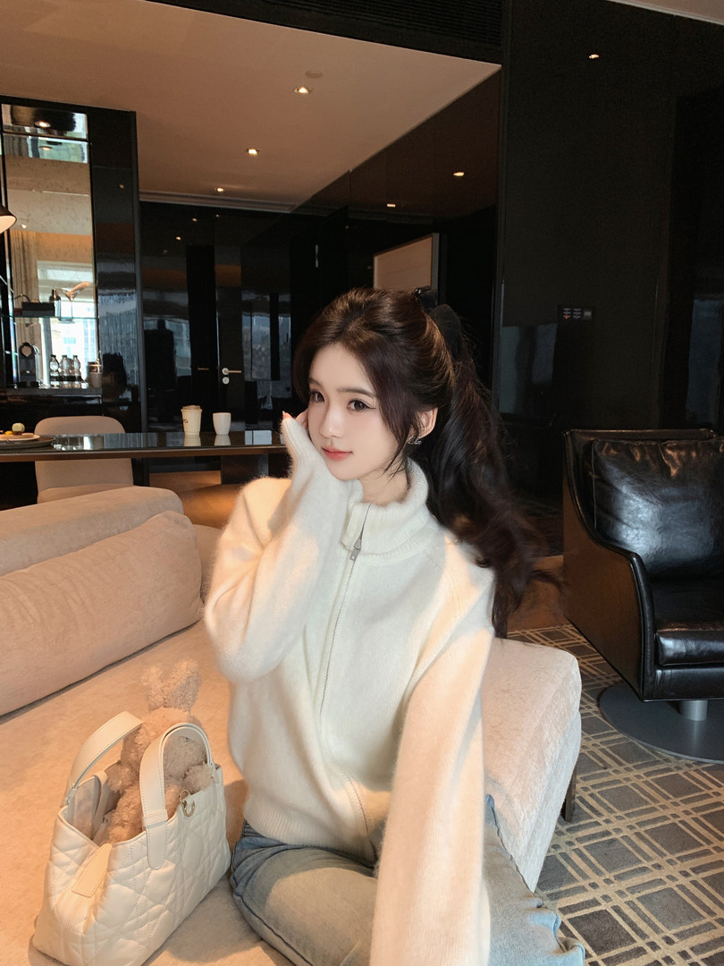 Woman wearing a white zip-up sweater, sitting on a couch with a white quilted handbag beside her.