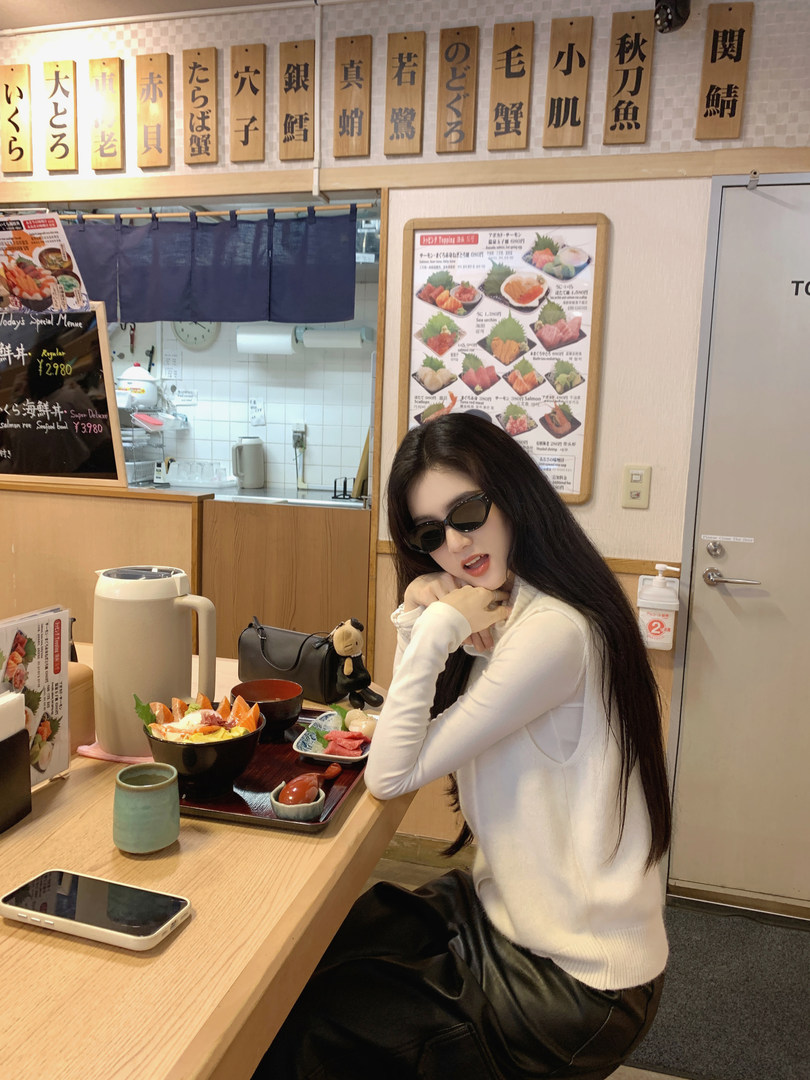 Woman wearing white long-sleeve top and black pants sitting at a sushi restaurant table with food and Japanese signage in background