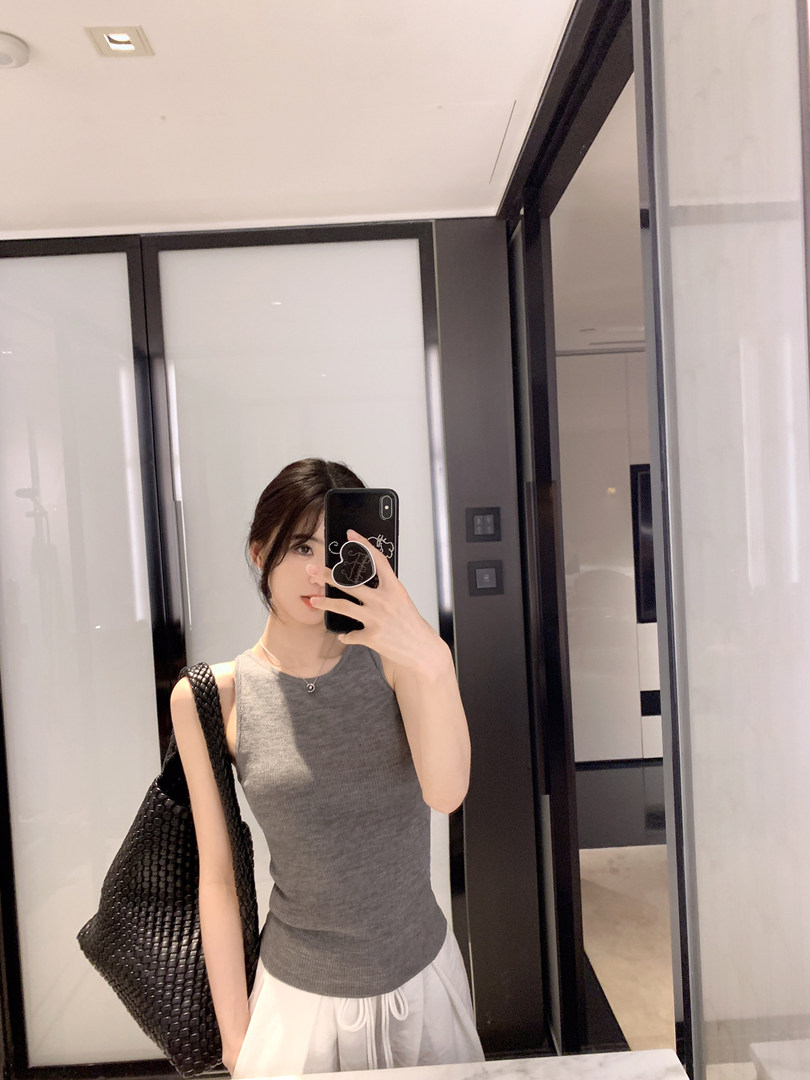 Woman taking mirror selfie wearing gray sleeveless top and white pants with black woven bag