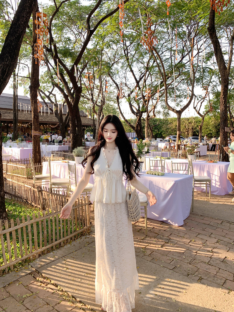 Woman wearing a white lace two-piece outfit outdoors at an event with decorated trees and tables
