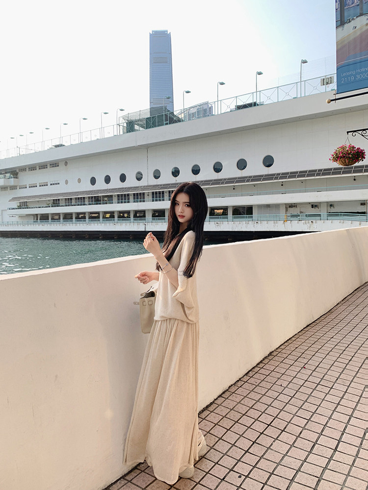 Woman wearing a beige long dress and matching top, standing by a waterfront with a building in the background.