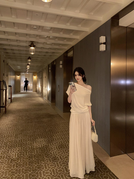 Woman wearing an off-shoulder white top and pleated maxi skirt in a hotel hallway