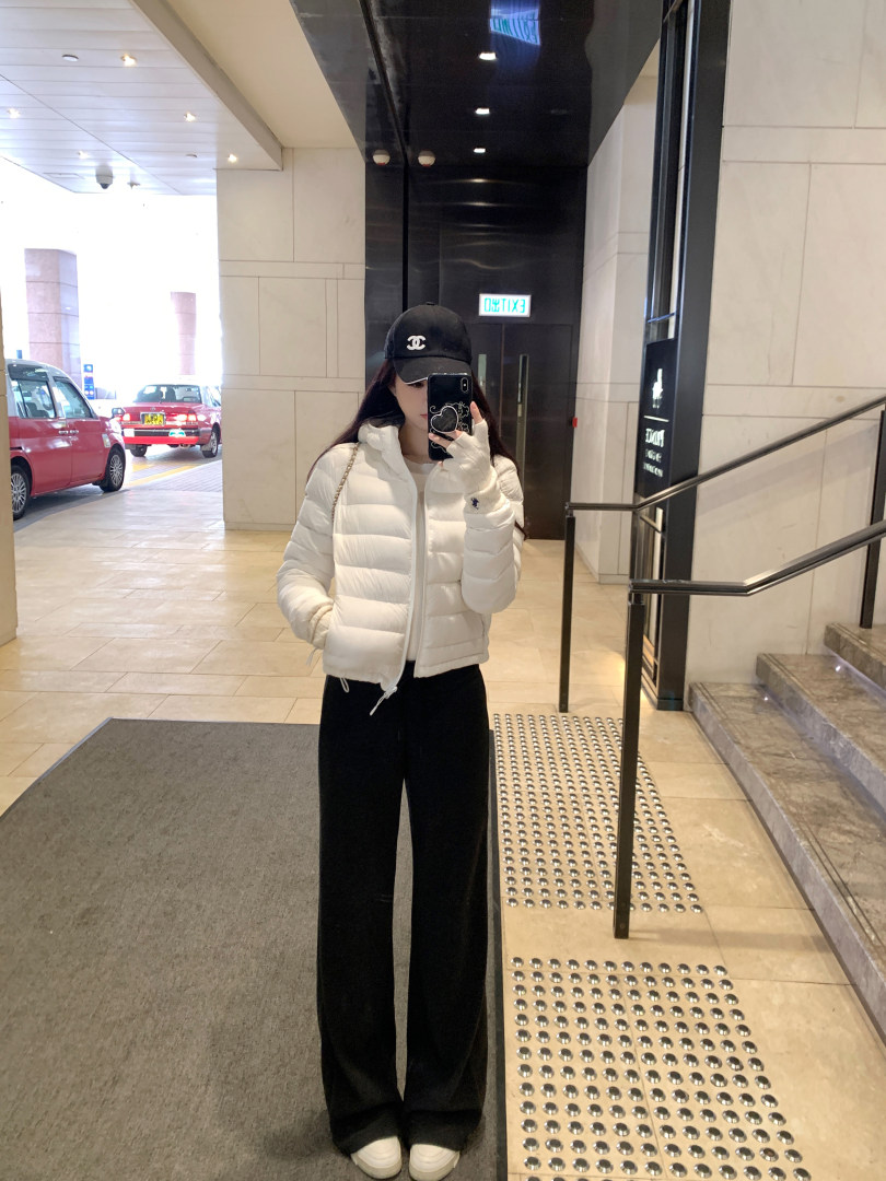 Person wearing a white puffer jacket, black pants, and a black cap taking a mirror selfie in a building lobby.