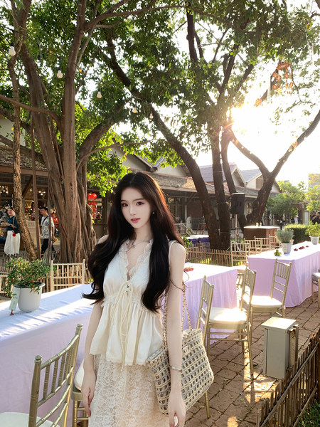 Woman wearing a white lace top and skirt with a woven bag, standing outdoors near tables and trees.