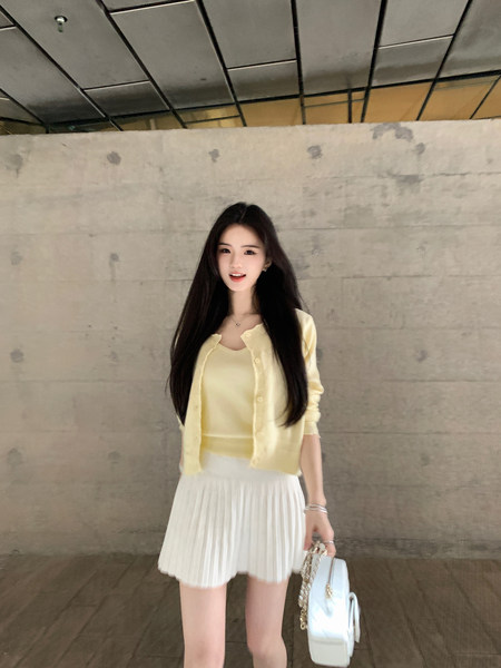 Woman wearing a light yellow cardigan over a matching top with a white pleated skirt, holding a white handbag.