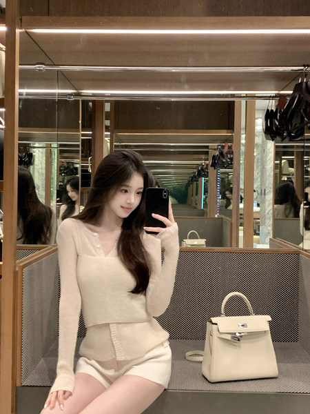 Woman taking mirror selfie wearing a beige knit top and matching shorts with a handbag beside her