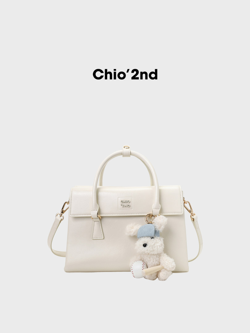 Cream handbag with top handles, shoulder strap, and teddy bear charm on a white background