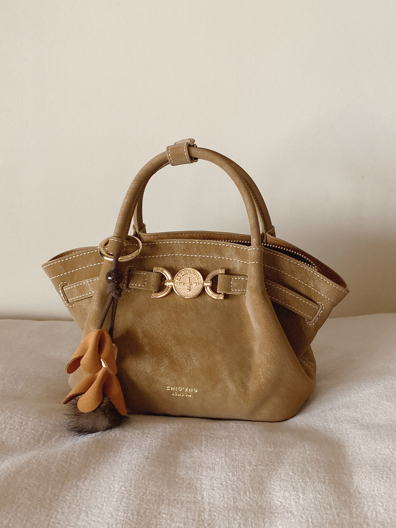 Tan leather handbag with top handles and decorative charm on a neutral background