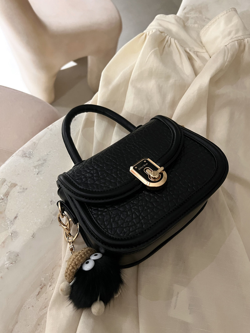 Black textured handbag with gold hardware and a fuzzy charm on a fabric backdrop