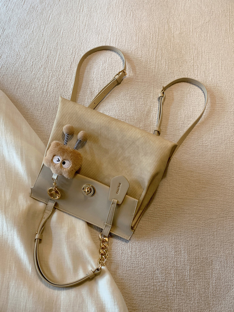 Beige handbag with long straps and a fuzzy bear charm laid on a textured fabric surface