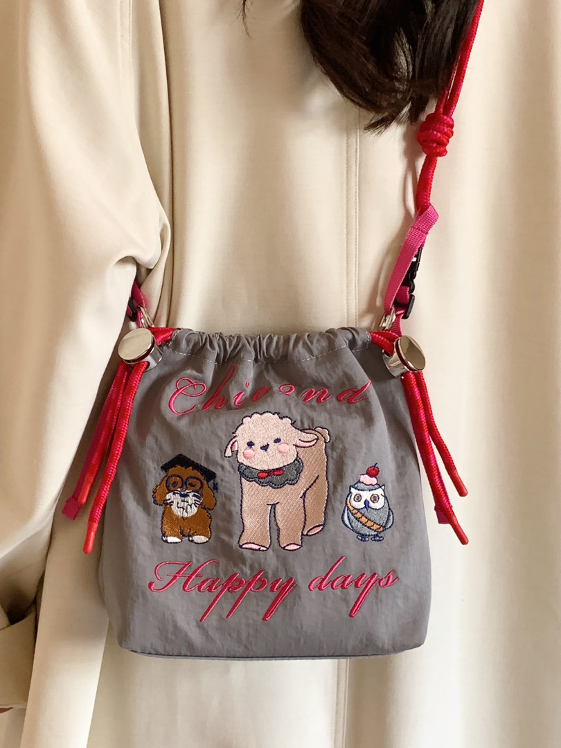 Gray drawstring bag with pink cords and embroidered animal design worn over a cream coat