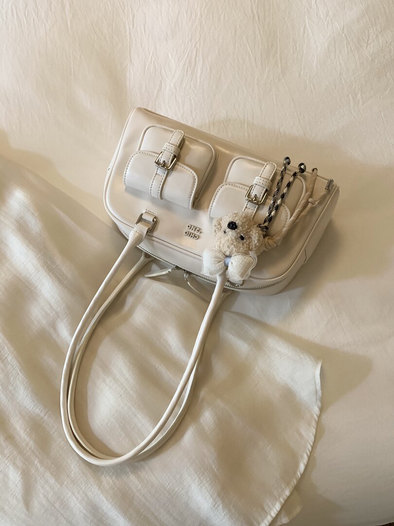 White leather shoulder bag with buckle details and a teddy bear charm on a soft fabric background