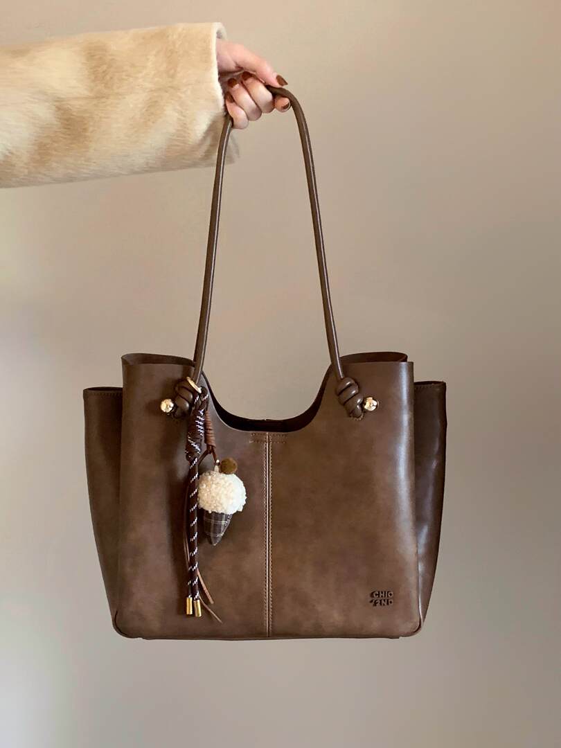 Brown leather tote handbag with long shoulder straps and charm detail being held by hand