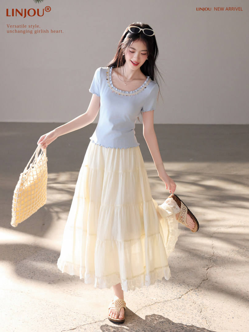 Model wearing a light blue top with a cream maxi skirt and woven sandals, holding a net bag