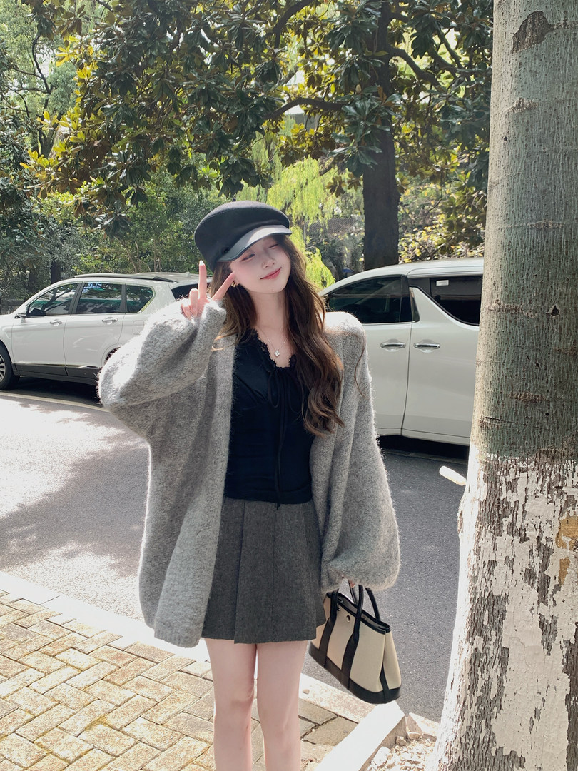Woman wearing a gray oversized cardigan and pleated skirt with a black cap and tote bag outdoors