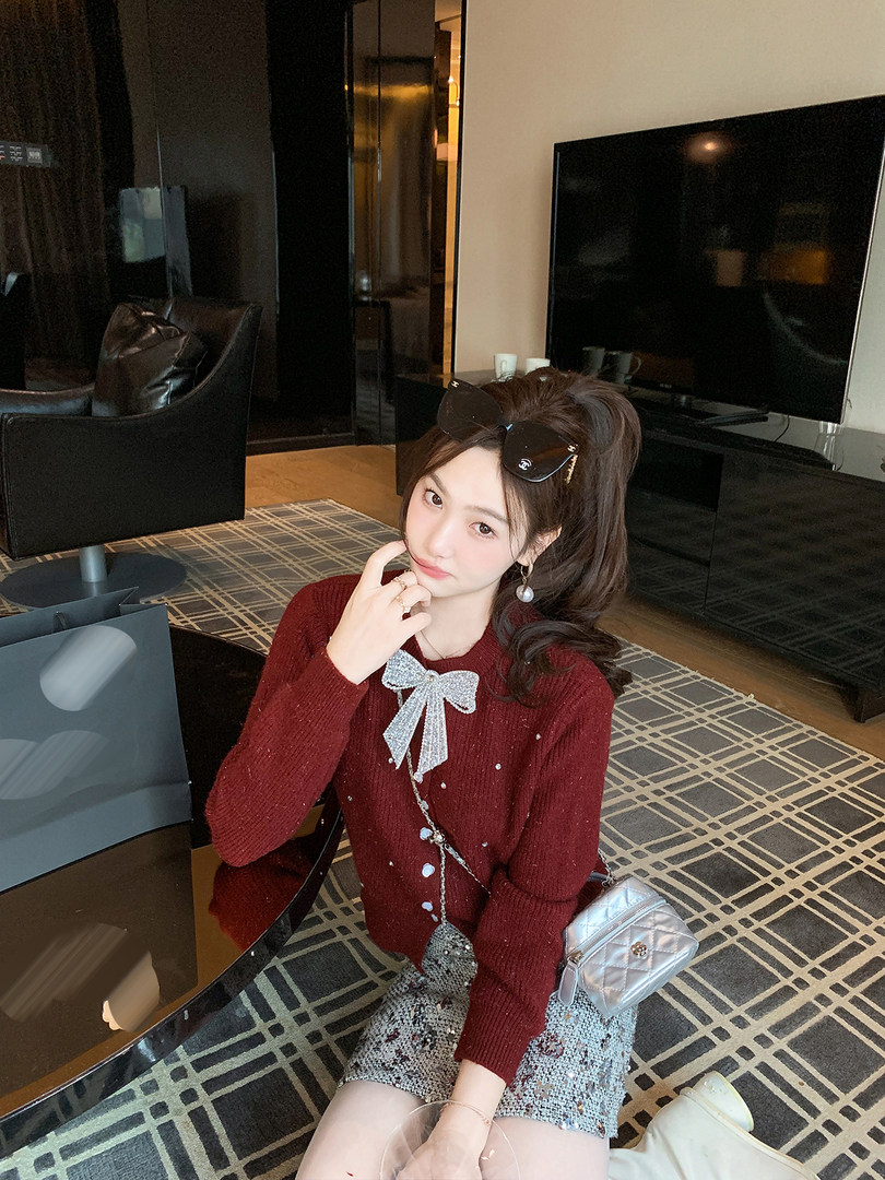 Woman wearing a burgundy knit sweater with a decorative bow and patterned skirt, posed indoors with a small quilted bag