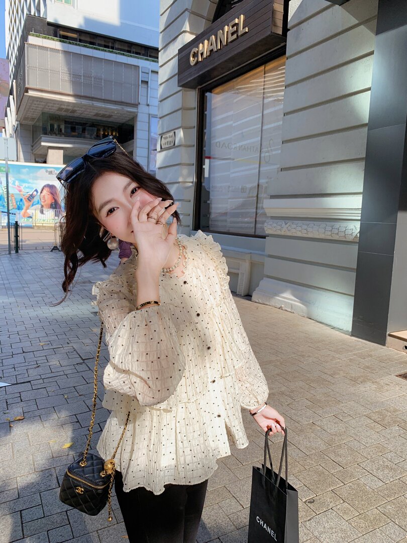 Woman wearing a cream ruffled blouse and black pants holding a Chanel shopping bag