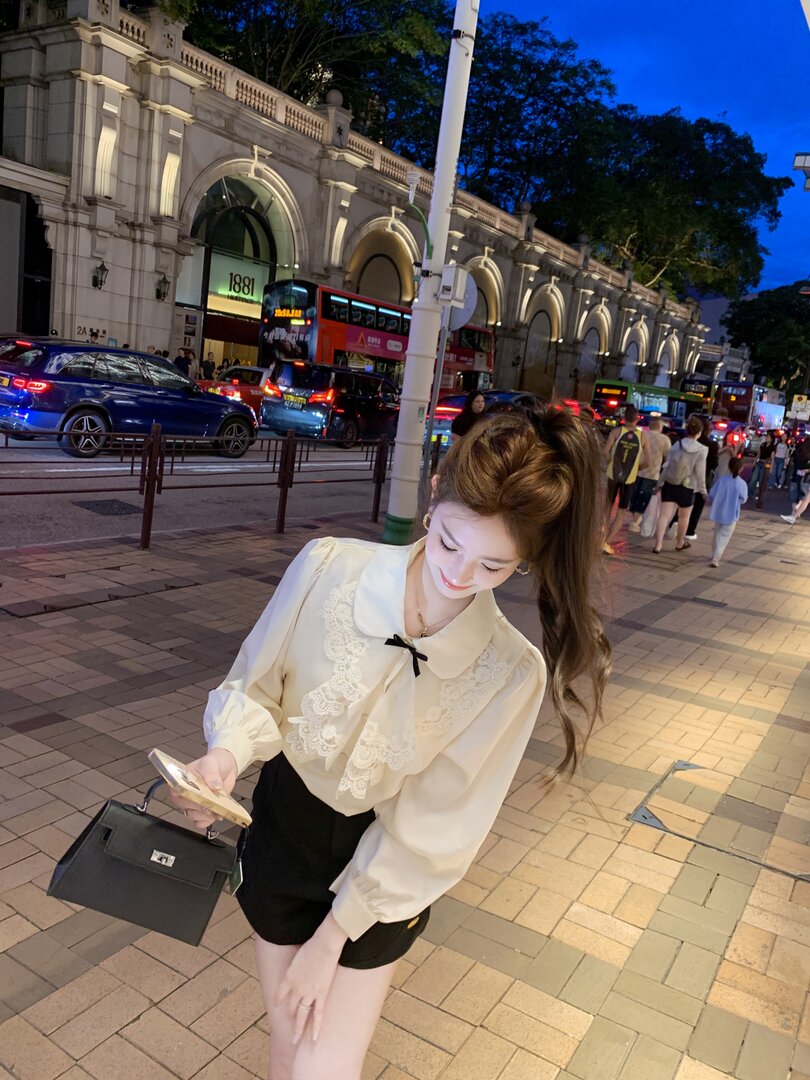 Woman wearing a cream lace-trim blouse with black shorts and a black handbag on a city street at night