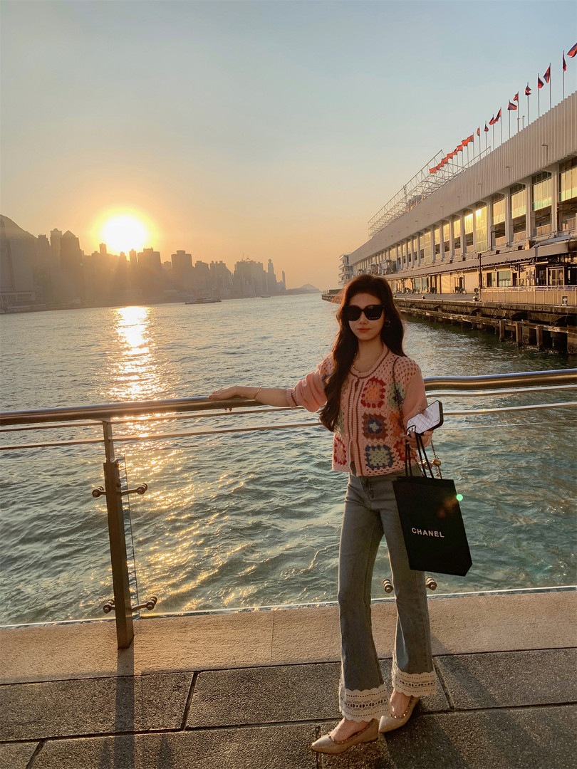 Woman wearing a crochet cardigan and flared jeans while holding a Chanel shopping bag by the waterfront at sunset