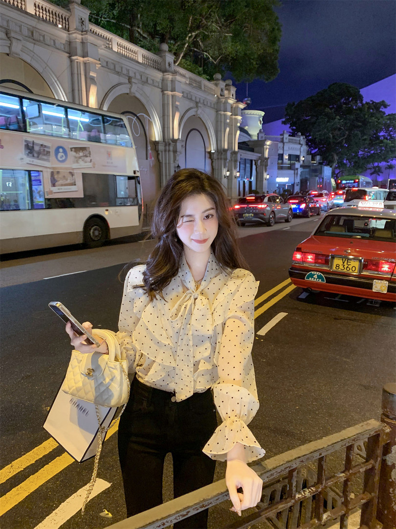 Woman wearing a sheer polka dot blouse and black pants with a white handbag at night